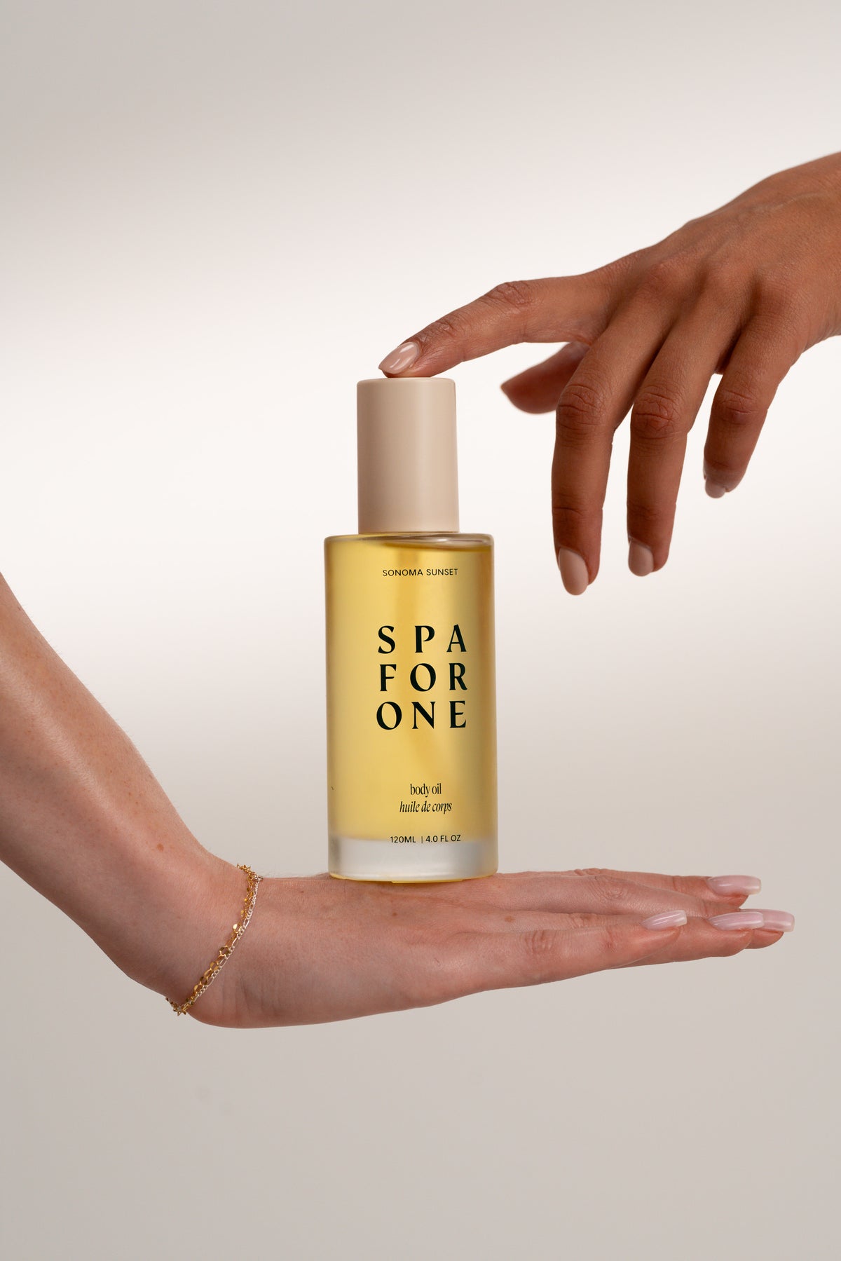 A bottle of Spa For One body oil is held between two hands against a soft beige studio background. The bottle is filled with golden-yellow oil and has a beige cap, with the brand name 'SPA FOR ONE' printed in black on the front.