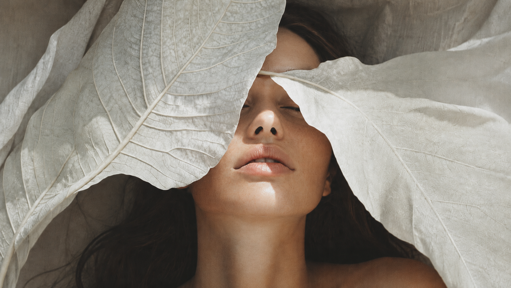 A close-up lifestyle shot of a woman lying down with large, textured off-white leaves draped over her face and eyes, with soft natural light highlighting her skin and the leaf textures.