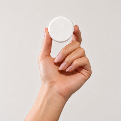 A hand with pale pink manicured nails holds a white shower steamer tablet against a clean, light gray background.