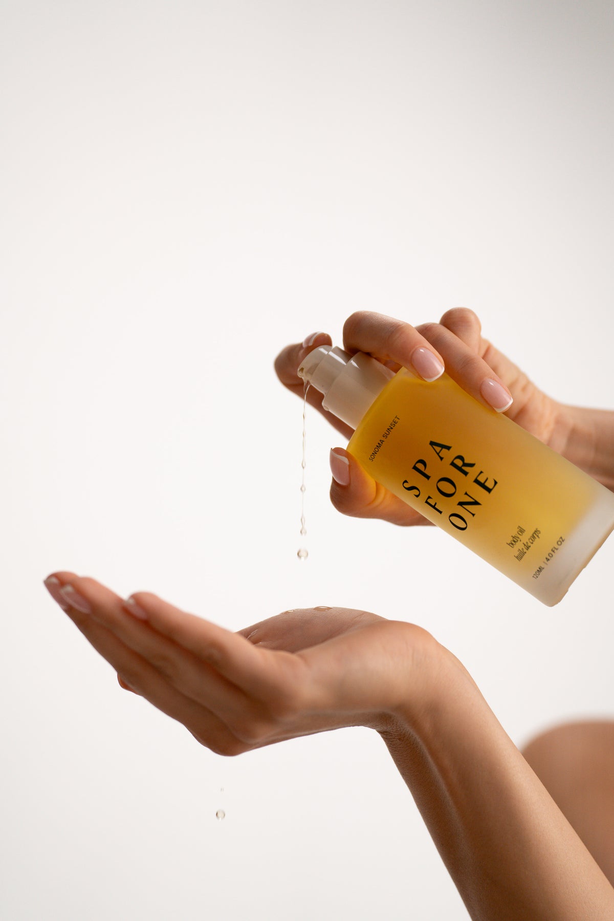 Hand pumping Sonoma Sunset body oil from a frosted glass pump bottle, a drop falling into an open palm