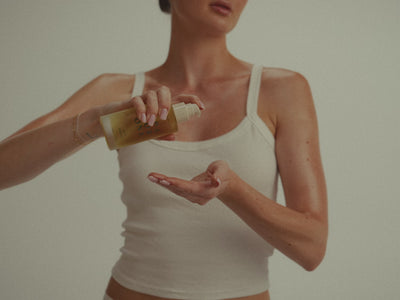 Woman in a white tank top pumping Spa For One body oil into her hand after the shower